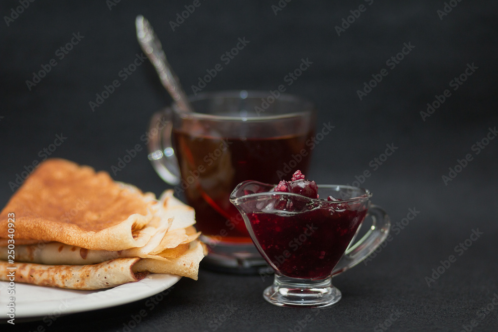 Tea with raspberry jam and pancakes. Cup of transparent glass, sauce-boat and white plate. Dark food photo style. Tea with raspberry jam and pancakes. Cup of transparent glass, sauce-boat and white plate. Dark food photo style.