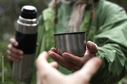 Obraz na plátne man in green jacket offers a hot drink - tea or coffee from thermos to someone w