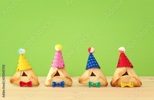 Purim celebration concept (jewish carnival holiday). Traditional hamantaschen cookies with cute clown hats over wooden table and green background.
