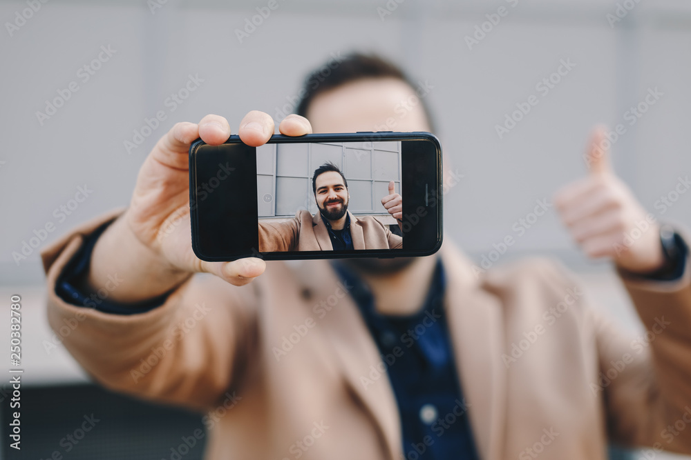 Close-up portrait of attractive and modern young man taking a selfie with his mobile phone, wearing fashionable clothes and posing for friends on social media. Focus on smartphone screen.