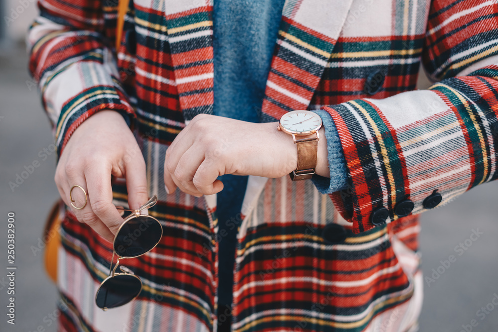 Close-up detail of fashion accessories, stylish young woman wearing an overcoat with a tartan pattern, a golden ring, a pair of modern sunglasses and a wrist watch.