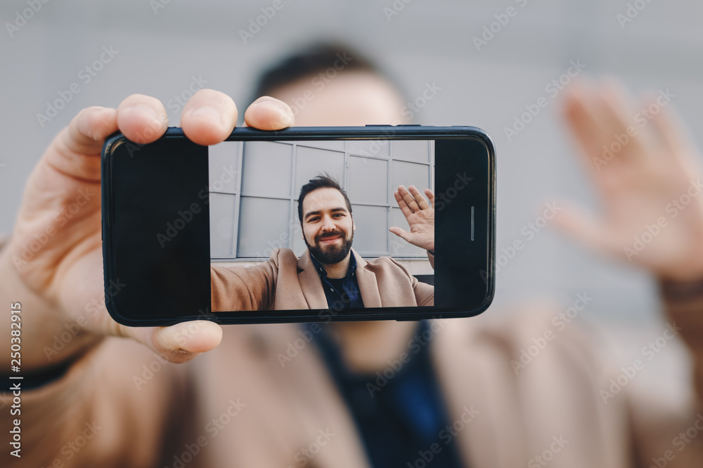 Close-up portrait of attractive and modern young man taking a selfie with his mobile phone, posing and waving for social media friends. Focus on device.