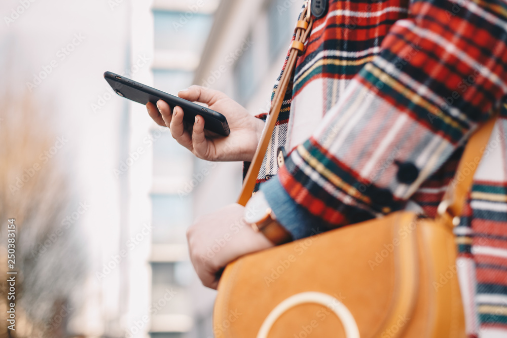 Close up detail of modern day female accessories - stylish young woman wearing an overcoat with a tartan pattern and a wrist watch while holding a fancy yellow bag and typing on her mobile phone.