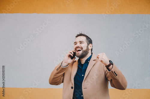 Portrait of attractive and happy young male freelancer clenching his fist in the middle of a phone conversation, celebrating good news that he received. Space for text and advertising. photo