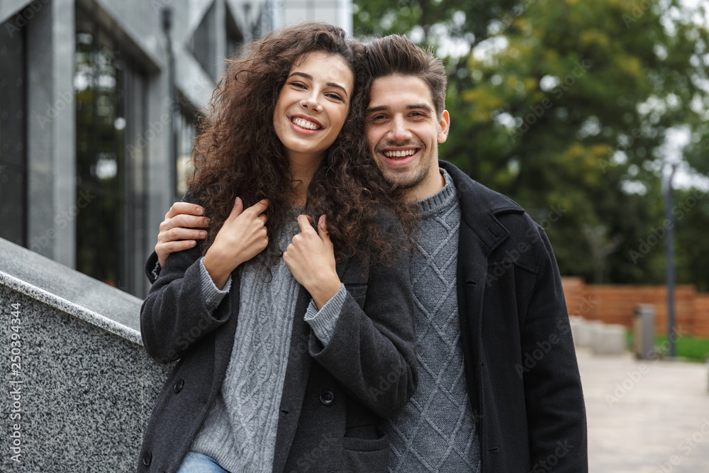 Image of joyous couple man and woman 20s in warm clothes, standing over gray building outdoor