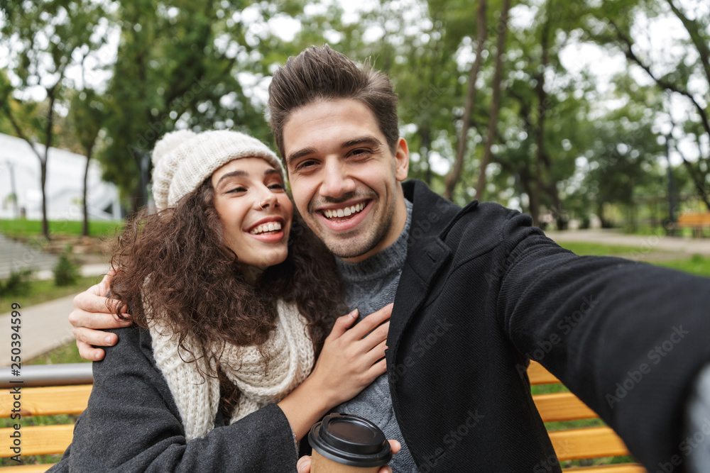 Portrait of amazing couple man and woman 20s taking selfie photo, while sitting on bench in green park Portrait of amazing couple man and woman 20s taking selfie photo, while sitting on bench in green park
