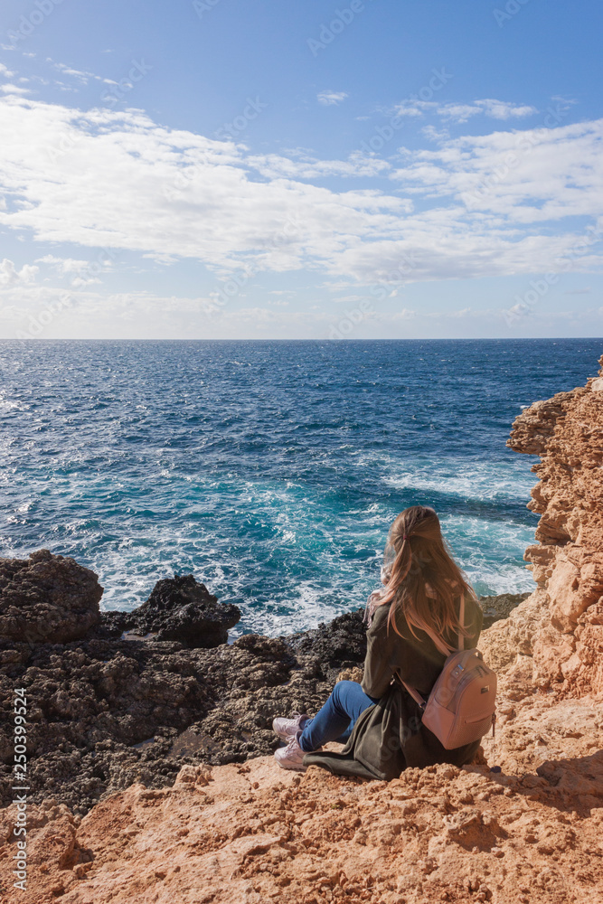 Happy female tourist in coat sits on cliff and enjoys sea view in Malta