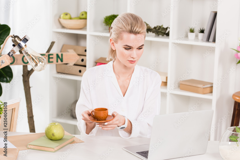attractive woman looking at laptop while holding cup in office attractive woman looking at laptop while holding cup in office