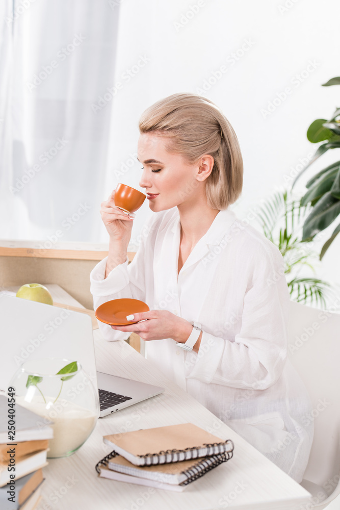 attractive woman drinking coffee from cup near laptop in office attractive woman drinking coffee from cup near laptop in office