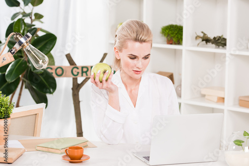 attractive woman looking at laptop and holding green apple in office