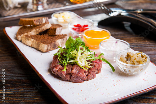 Beef tartare with arugula salad, crisp bread chips, sauces and snacks on white plate on the served restaurant table.
