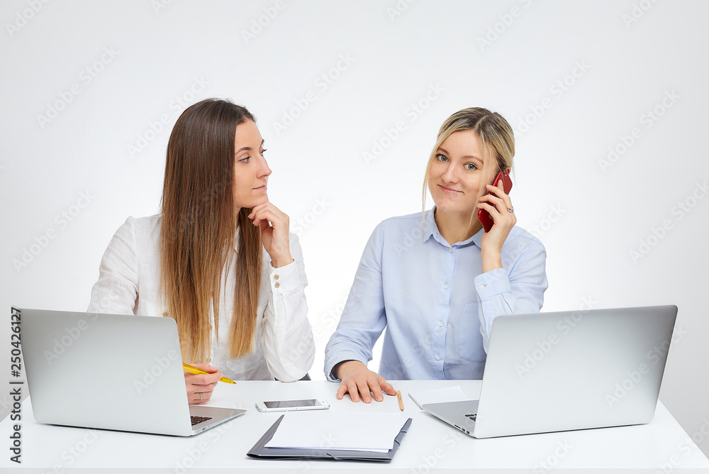 Happy blonde caucasian female manager calls by the cell phone nearby her brunette female colleague in the office with the white background. Happy blonde caucasian female manager calls by the cell phone nearby her brunette female colleague in the office with the white background.
