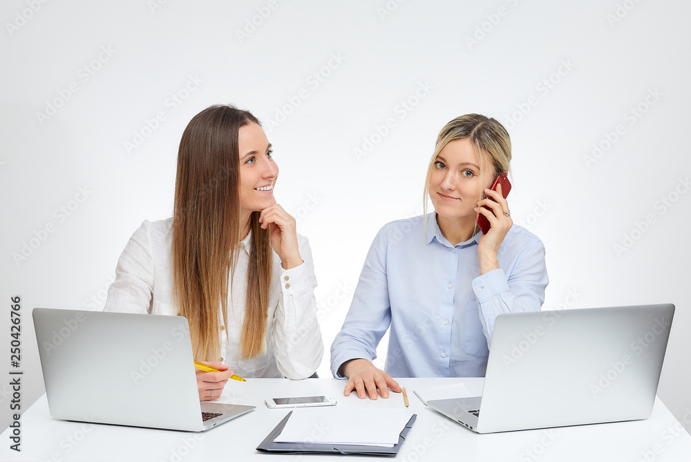 Happy blonde caucasian female manager calls by the cell phone nearby her brunette female colleague with a smile in the office with the white background. Happy blonde caucasian female manager calls by the cell phone nearby her brunette female colleague with a smile in the office with the white background.
