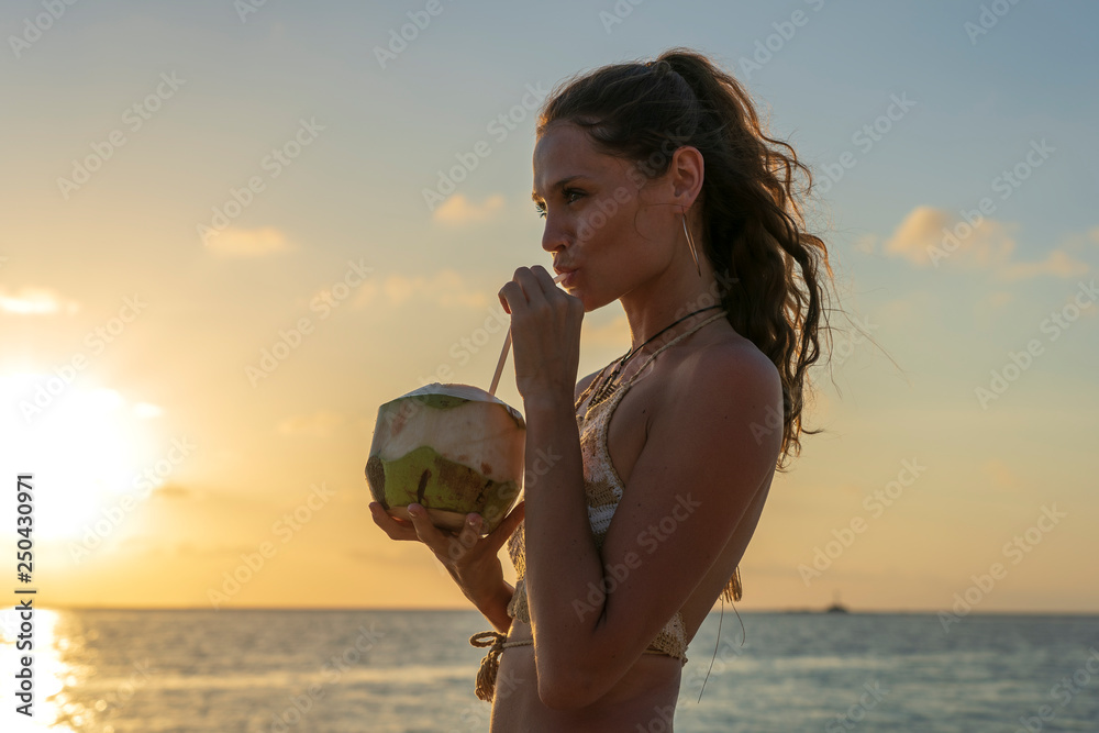 Young beauty girl drinking coconut at tropical beach near sea water at paradise island at sunset. Summer concept. Holiday travel.