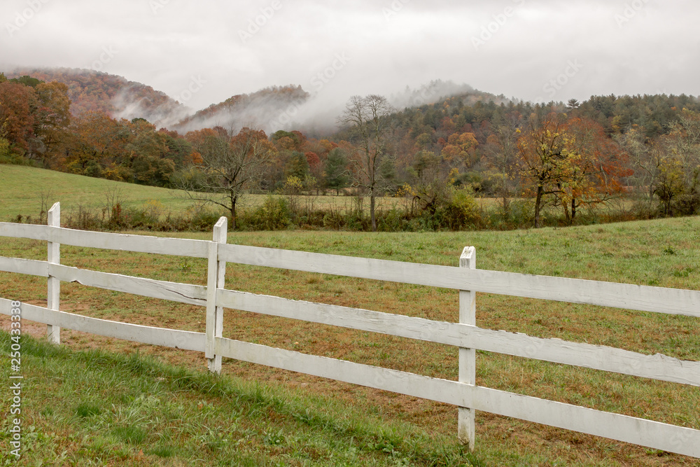 White fence in front of green pasture with fall colored trees and stratus clouds