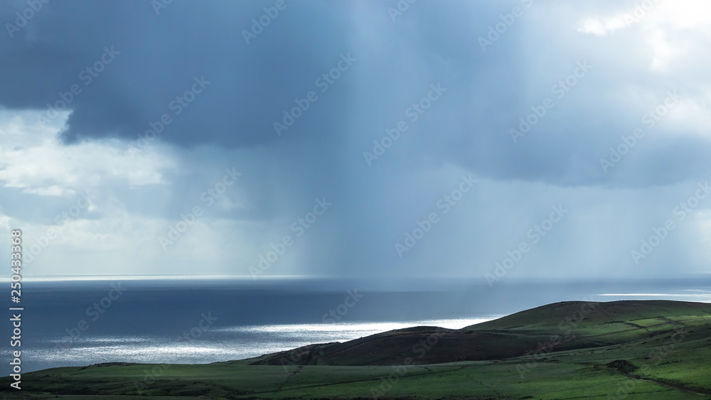 Heavy rain over calm ocean in the background. Green cliff in foreground.