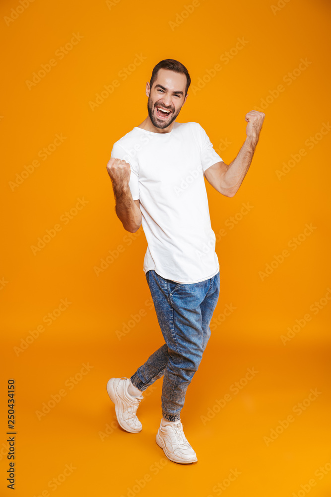 Full length image of optimistic guy 30s in t-shirt and jeans clenching fists while standing, isolated over yellow background Full length image of optimistic guy 30s in t-shirt and jeans clenching fists while standing, isolated over yellow background