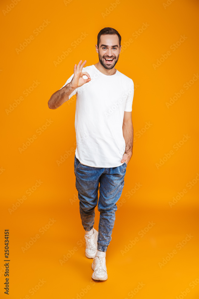 Full length image of handsome guy 30s in t-shirt and jeans showing ok sign while standing, isolated over yellow background Full length image of handsome guy 30s in t-shirt and jeans showing ok sign while standing, isolated over yellow background
