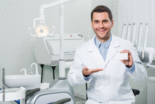 cheerful dentist in white coat smiling while showing blank card