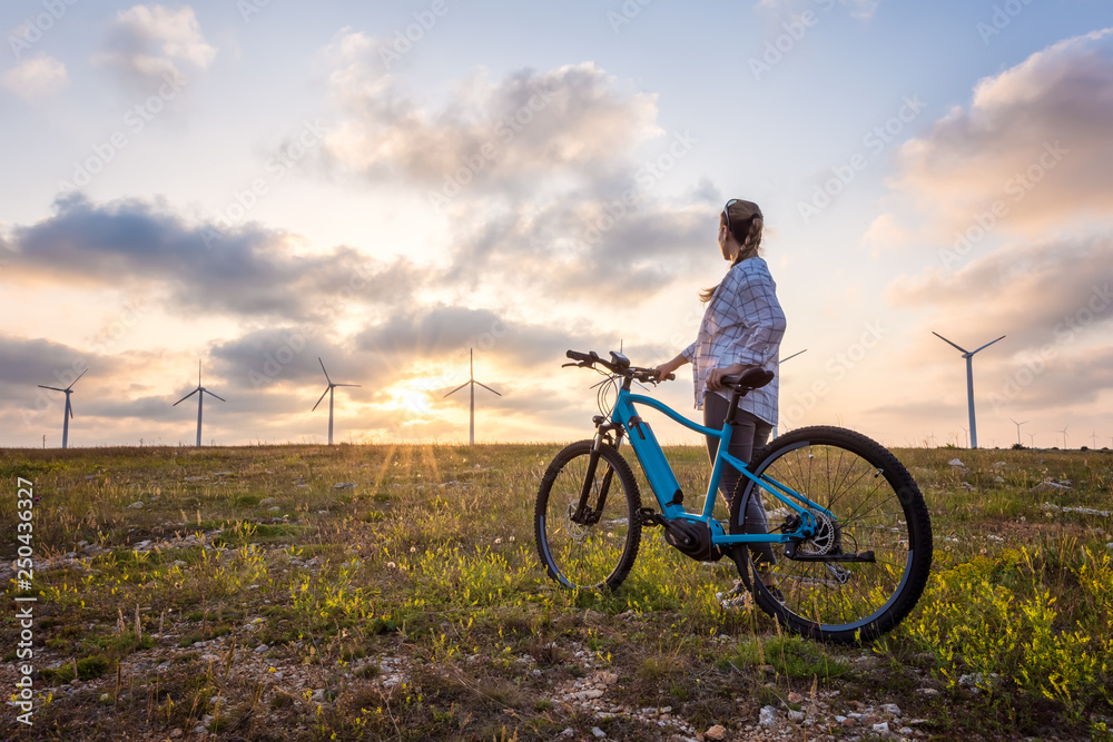 Woman with a bike in the nature / A woman with a bike enjoys the view of sunset over a summer field with a wind farm