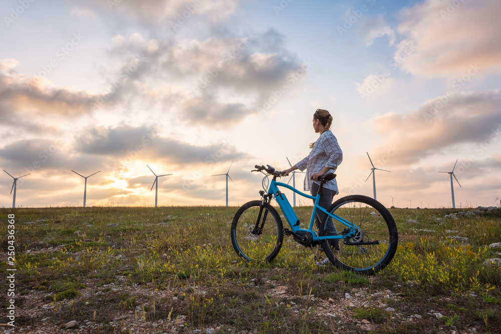 Fototapeta premium Woman with a bike in the nature / A woman with a bike enjoys the view of sunset over a summer field with a wind farm