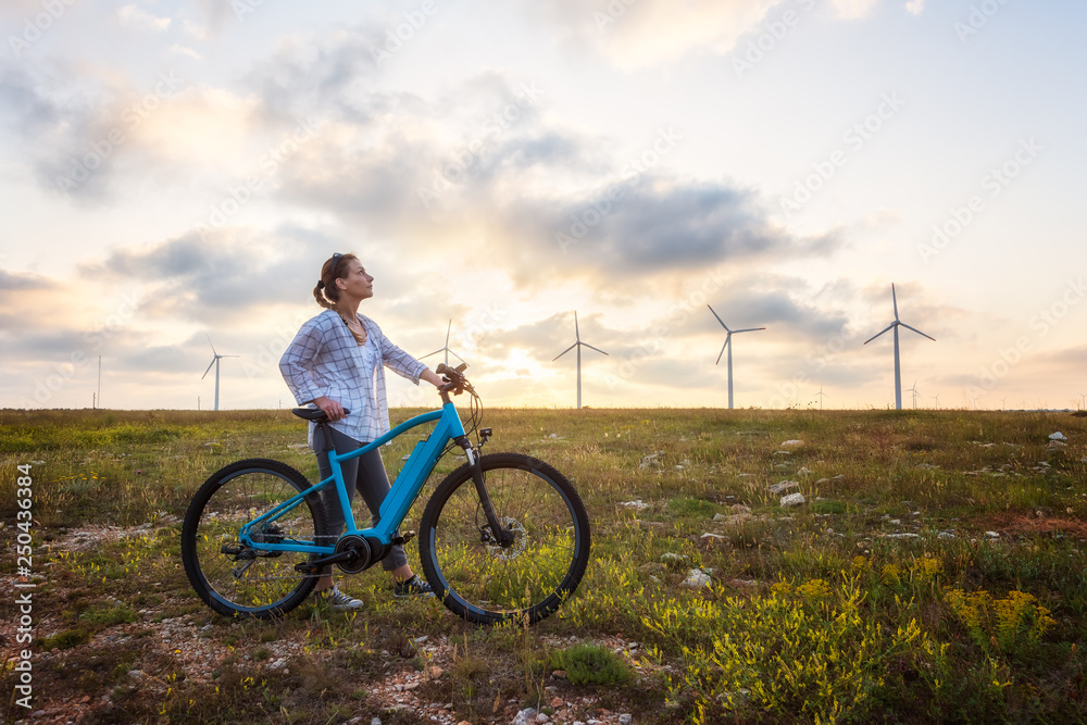Obraz premium Woman with a bike in the nature / A woman with a bike enjoys the view of sunset over a summer field with a wind farm