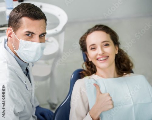 selective focus of dentist in mask near smiling patient showing thumb up