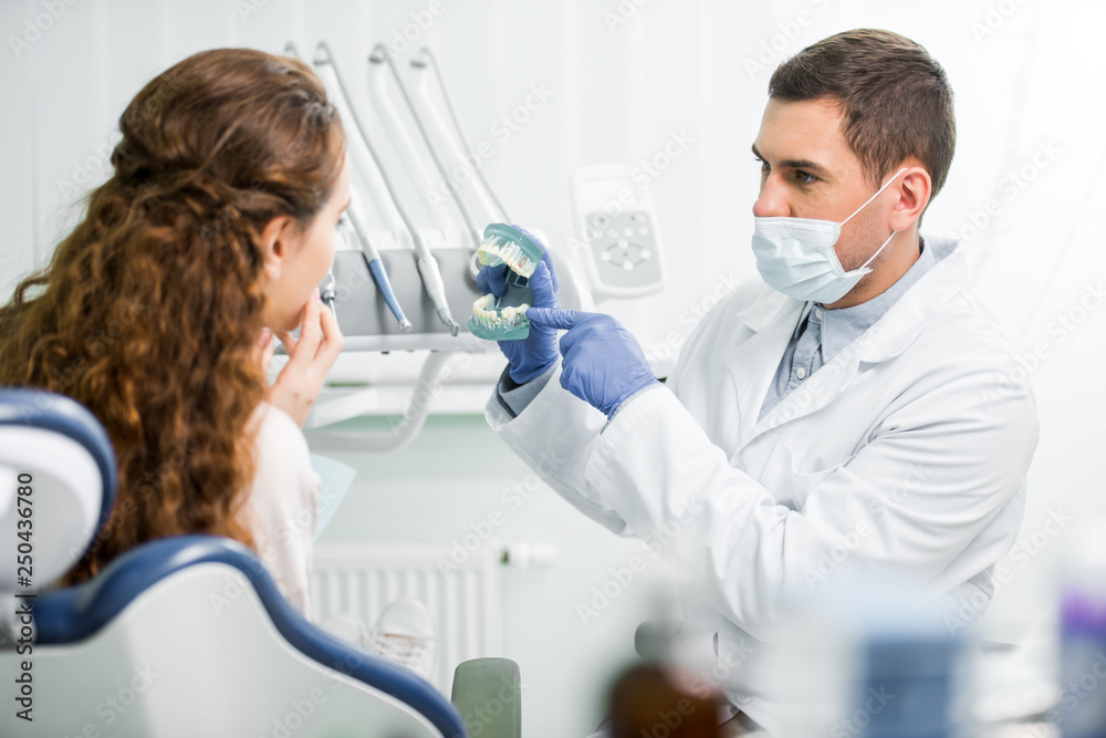 selective focus of dentist in mask and latex gloves pointing with finger at teeth model near female patient selective focus of dentist in mask and latex gloves pointing with finger at teeth model near female patient