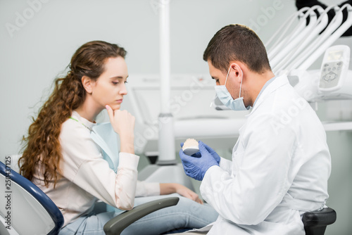 dentist in latex gloves and mask holding teeth model near pensive woman