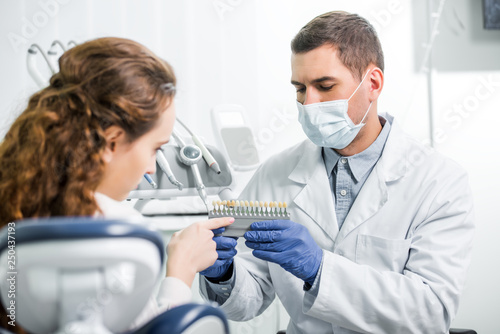 woman pointing with finger at teeth color palette near dentist in mask and latex gloves