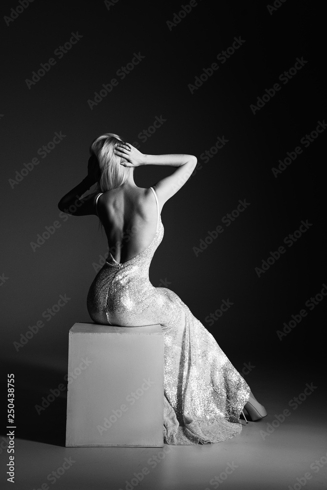 Fototapeta premium High fashion portrait of young elegant woman. Young lady in amazing silver glitter sequins luxury dress sitting on armchair Studio shot. Back view 