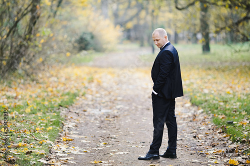 Handsome groom in classy black suit poses in autumn park covered with fallen leaves
