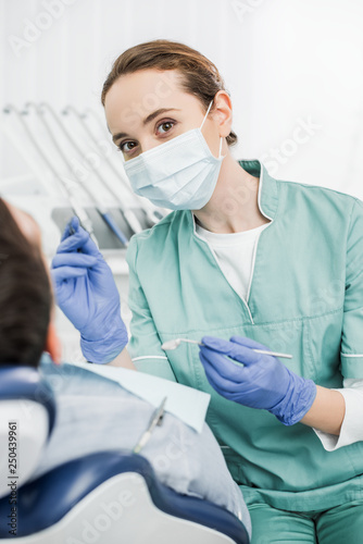selective focus of female dentist in mask with dental instruments in hands near patient