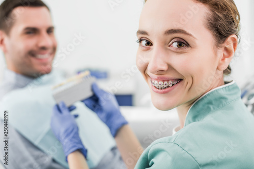 selective focus of cheerful dentist with braces on teeth smiling while holding color palette near patient