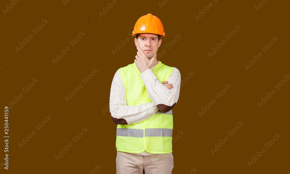 The thoughtful man in a construction helmet and a vest, looks in the camera, the crossed hands at the head
