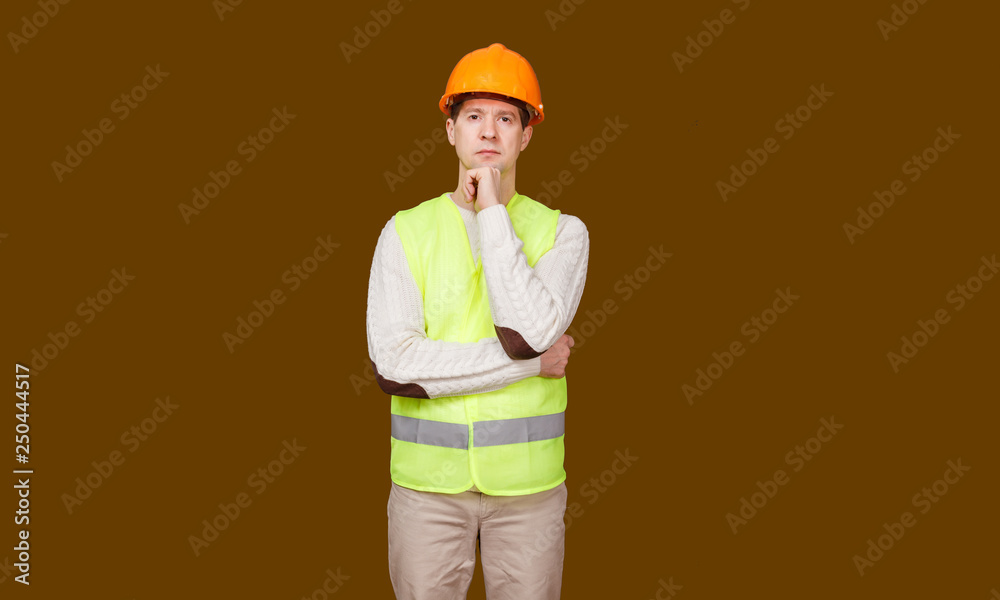 The serious man in a construction helmet and a vest, looks in the camera, the crossed hands at the head