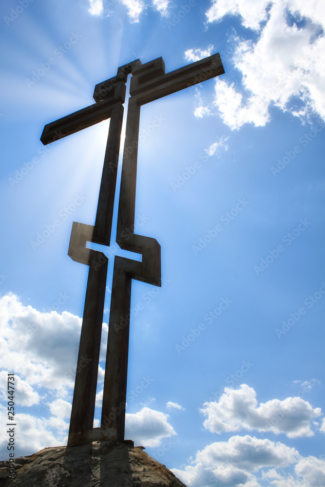 photo of a metal orthodox cross close-up in projection against a blue sky with clouds and a bright sun glare cross