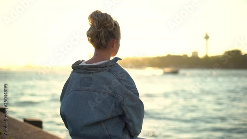 Back view of young pretty blond girl sitting on concrete pier enjoying warm sunset shine looking at boat floating along sea creating small waves. Woman daydreaming admiring cityscape near water
