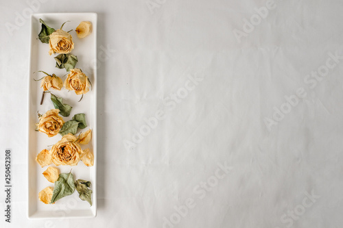 Dried yellow rose flower and dried leaves on white plate on white table top with fabric texture with empty copy space. Selective focus.