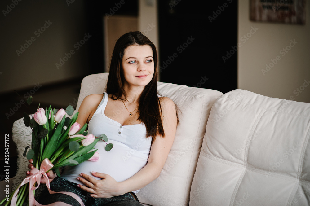 Pregnant happy woman sitting on bed and touching her belly holding a bouquet of flowers, hugging tummy at home. Baby Shower. Motherhood concept. Pregnancy, rest people, expectation concept. Pregnant happy woman sitting on bed and touching her belly holding a bouquet of flowers, hugging tummy at home. Baby Shower. Motherhood concept. Pregnancy, rest people, expectation concept.