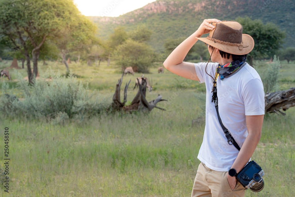 Young Asian man traveler and photographer holding hat in safari looking at wildlife animals. Travel safari in Africa concept Young Asian man traveler and photographer holding hat in safari looking at wildlife animals. Travel safari in Africa concept