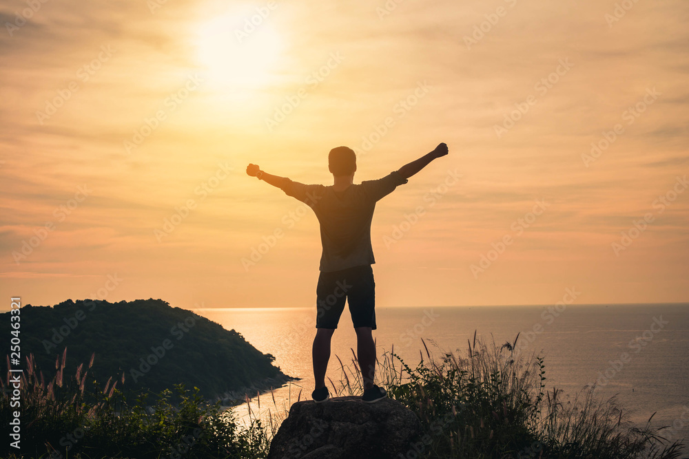 On the edge of a rocky cliff a man raises his hands to heaven as a sign of freedom or victory and in the sky. Man on mountain summit enjoying aerial view hands raised over clouds .