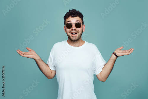 I don't now. Bearded man in sunglasses and white t sirt is gesturing unknown sign isolated over blue background. Young hipster with confuce face expression photo