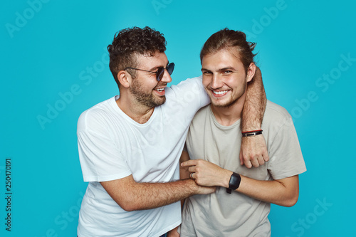 Image of ttwo cheerful handsome guys embracing each other, wearing casual clothes, isolated over blue studio background photo