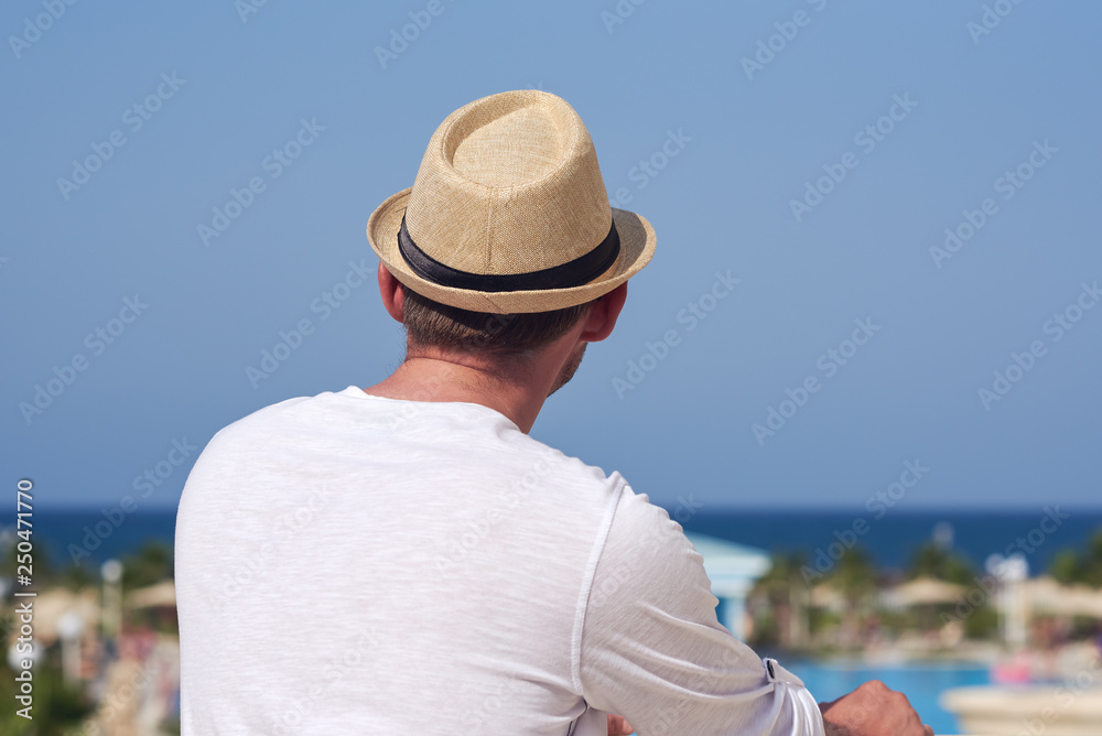 Man in sunhat relaxing against blue sea and enjoying picturesque luxurious hotel’s view.