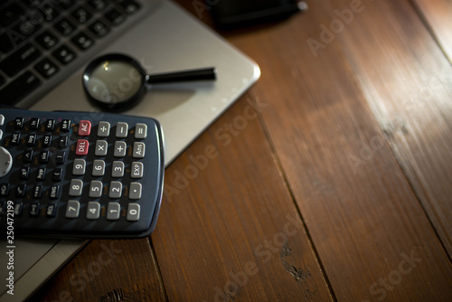 Magnifying glass on laptop computer, calculator, entering light, wooden background, space for text.