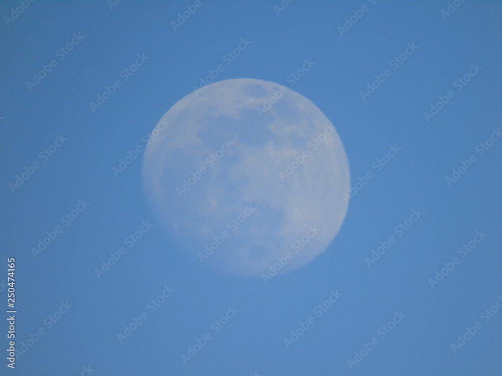 Liguria, Italy - 02/18/2019: An amazing photography of the full moonlight over the mountains, hidden and out of the trees in the village by day with beautiful blue sky in the background in winter days Liguria, Italy - 02/18/2019: An amazing photography of the full moonlight over the mountains, hidden and out of the trees in the village by day with beautiful blue sky in the background in winter days