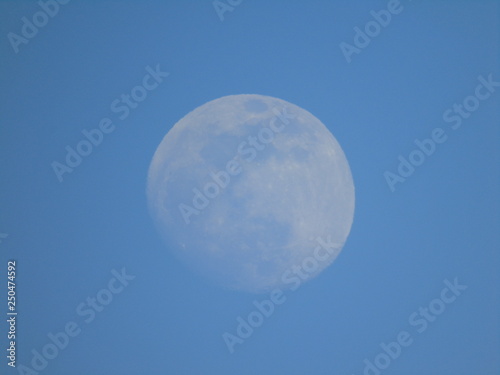 Liguria, Italy - 02/18/2019: An amazing photography of the full moonlight over the mountains, hidden and out of the trees in the village by day with beautiful blue sky in the background in winter days