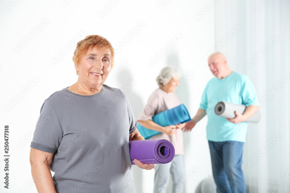 Elderly woman with yoga mat indoors. Space for text