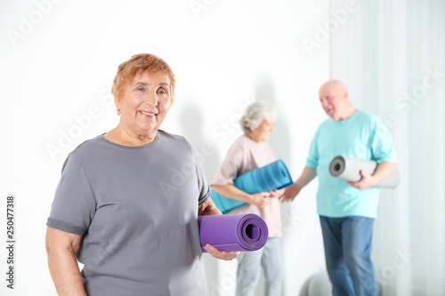 Elderly woman with yoga mat indoors. Space for text